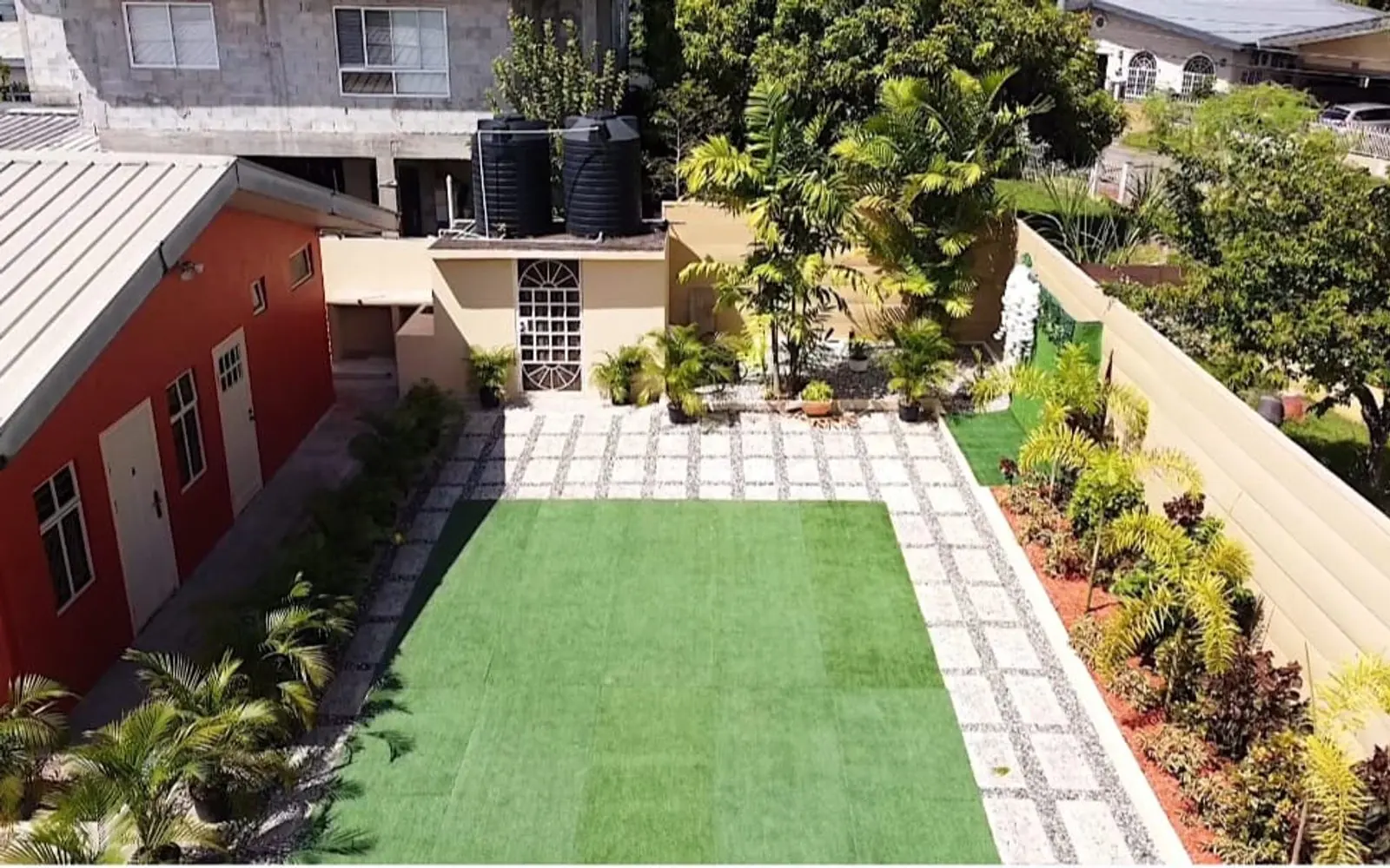 Aerial view of the G.E.M.S De La Rosa garden: a green lawn framed by palms, paved walkways, and a red-walled guest house